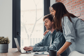 Naklejka premium Happy young family couple bonding laughing using laptop looking at screen sit at table, smiling husband and wife buying choosing goods online doing internet shopping order with delivery at home.