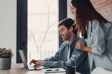 Happy young family couple bonding laughing using laptop looking at screen sit at table, smiling husband and wife buying choosing goods online doing internet shopping order with delivery at home.