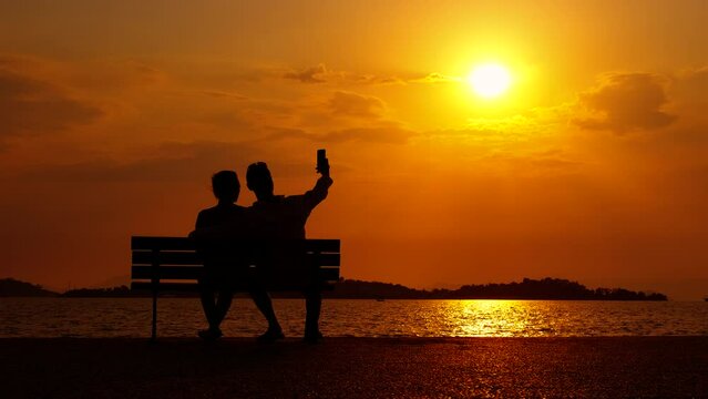 Loving family silhouette with phone at dusk. A view of lovely couple silhouette takes photo on the bench during sea dusk in summer.
