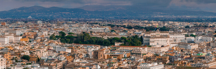 Obraz premium An aerial view of Rome shows a dense cityscape, terracotta roofs, and mixed architecture under a cloudy sky. Its rich history is seen in the mix of old and new buildings, nestled among hills.