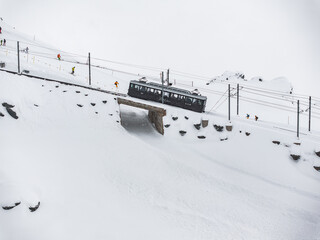 A snowy Zermatt, Switzerland from above, with a dark cogwheel train on a snow bridge, encircled by electrical lines. Skiers in bright gear on a gentle slope showcase winter sports and alpine travel.