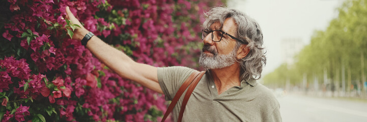 Friendly middle aged man with gray hair and beard wearing casual clothes, wall with purple flowers...