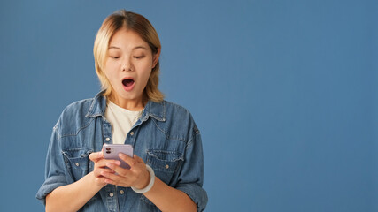 Surprised young woman dressed in denim shirt, with mobile phone in hands getting good news, mockup...