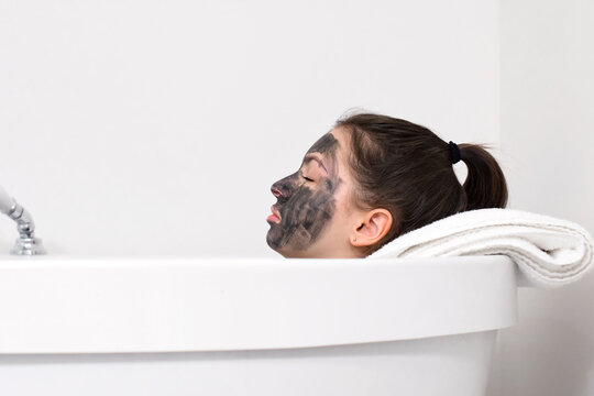 Young girl lying inside bathtub with mud face mask resting head on towel