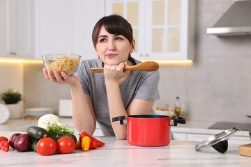 Pensive housewife with spoon and raw pasta at white marble table in kitchen. Cooking process
