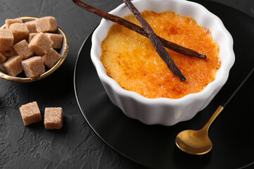 Delicious creme brulee in bowl, vanilla pods, sugar cubes and spoon on dark gray textured table, closeup