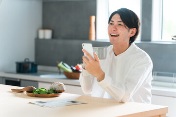 ダイニングで朝食を取りながらスマホを見る若いアジア人男性
