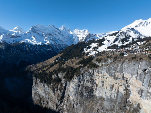 An aerial view of Murren, Switzerland, reveals snow covered chalets on a slope and buildings near a cliff leading to a gorge, all against snow capped peaks and a clear blue sky.