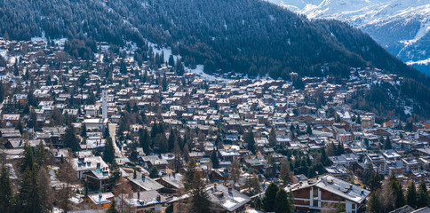 Aerial view of Verbier, Switzerland, showcasing snow covered chalets and buildings typical of alpine architecture, nestled in a forested mountain landscape with the Swiss Alps in the background.