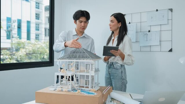 Beautiful young caucasian project manager brief asian engineer by using tablet while engineer measuring house model on meeting table with blueprint, architectural equipment scatter around. Immaculate