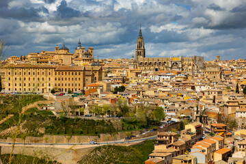 Scenic Toledo cityscape on hill above Tagus river with view of tall bell tower of medieval gothic Roman Catholic cathedral and domes of Church of San Ildefonso against cloudy sky in spring day, Spain