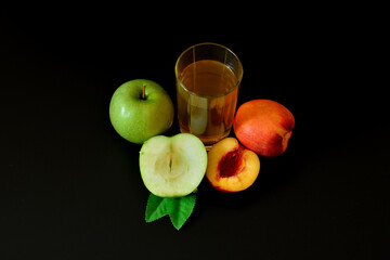 A glass of fruit juice on a black background, next to pieces of a ripe peach and a green apple with leaves.
