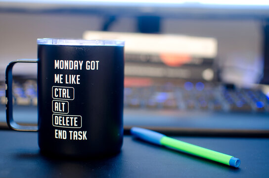 Coffee cup with the phrase "Monday got me like CRTL + ALT + DELETE END TASK" with a green-blue pen in a black table . Books , computer monitor and a keyboard in the background.