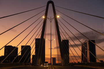 Sunrise on modern symmetrical suspension bridge with illumination, and road traffic. New high-rise buildings fill the background.