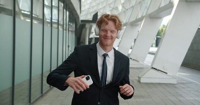 Entrepreneur in suit near office and plays cheerful music on phone through headphones. Formal suit contrasts with relaxed dance to music that brings positivity. Music helps brighten up busy day work.