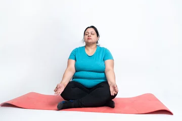 Fototapeten Overweight fat indian sitting on yoga mat doing yoga or meditation with hands in lotus postion. isolated over white background. Plus size female. copy space © gajendra