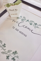 A white wedding welcome sign adorned with green floral designs, accompanied by a thank you note.