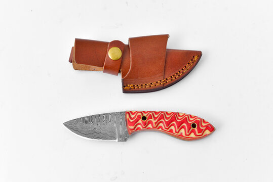 A small Damascus bladed knife with big red orange handle and leather sheath against a white background.