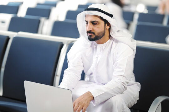 Emirati UAE Dubai man at airport ideal for business trip travel concept. Arab in Kandura dish dasha ghutra. Middle Eastern working using laptop while on the go