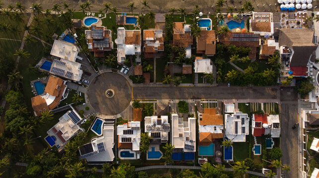 vista a&eacute;rea de casas de playa junto a la arena y el mar con sombrillas playeras techos de casas con alberca casas sobre la avenida con glorieta llena de &aacute;rboles en la costa