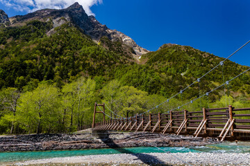 Wooden suspension bridge over a clear river surrounded by forest and mountains