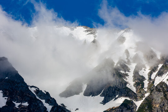 Clouds over the top of tall, jagged, snow covered mountain peaks