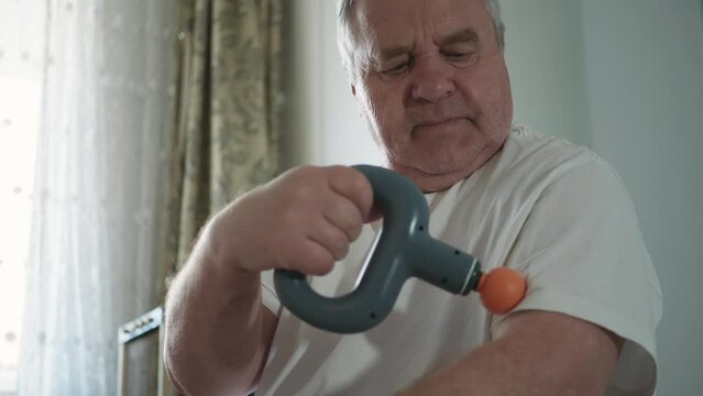 Elderly man massaging himself with massage gun at home. Electric vibration device for percussive therapy soft tissue massage that uses vibrations to relieve muscle tension.
