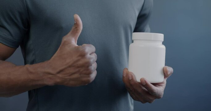 Close-up of male hands pointing at bottle of multivitamin with thumbs-up gesture while African American sportsman advertising supplements. Healthy nutrition and sports concept.