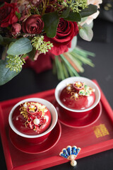 Two red cups filled with tea are adorned with golden jewelry and pearls, placed on a dark red tray inscribed with Chinese characters. A bouquet of deep red roses and green foliage adds to the scene
