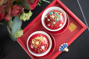 Two red cups filled with tea are adorned with golden jewelry and pearls, placed on a dark red tray inscribed with Chinese characters. A bouquet of deep red roses and green foliage adds to the scene