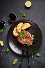 Tuna and avocado tartare with sesame seeds, capers and egg yolk on a dark ceramic plate with...