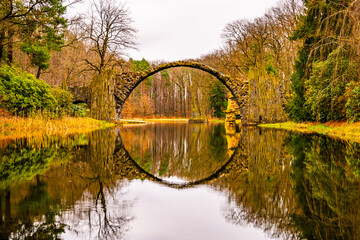 The Rakotzbrucke, also known as Devils Bridge, is reflected in calm waters on an overcast autumn day, surrounded by vibrant foliage. Germany