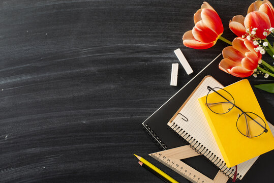 Embrace chic Teacher's Day theme. Top view snapshot of school supplies, notebooks, pencils, chalk, glasses, and vibrant tulip bouquet on chalkboard backdrop with space for greetings or promotions