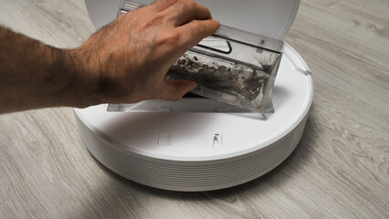 A hand empties a robot vacuum's dust collector into a trash can.