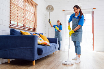 Two Asian young professional cleaning service women worker team working in the house. The girl housekeeper wiped a wet mop on wooden floor with another one clean the curtain with a feather duster.
