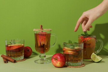 Glasses with apple cider, female hand, cinnamon sticks, piece of lime and red apples on green...