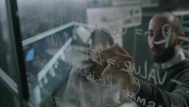 Through-glass footage of male and female alternative energy specialists doing calculations working with formulas while engineering new low-carbon technologies in office