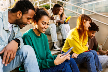 Group of young people using mobile phone sitting at staircase in the city. Millennial student friends having fun watching social media content on cell app. Technology lifestyle concept - obrazy, fototapety, plakaty