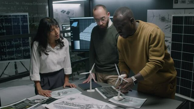 Side full medium footage of multiethnic team of alternative energy specialists discussing new ideas on wind turbines enhancement using windmill models, blueprints and drawings during office meeting