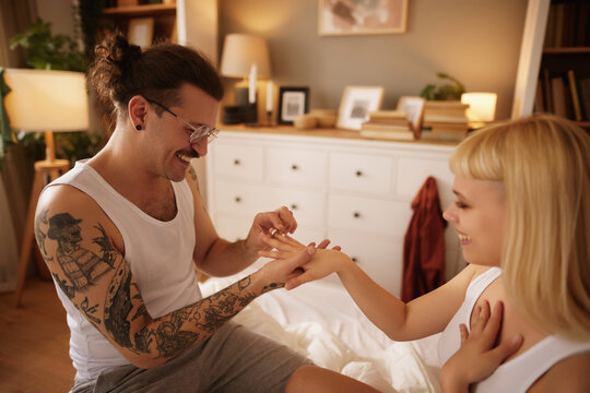 Tattooed man smiling while putting engagement ring on finger of delighted woman in bed
