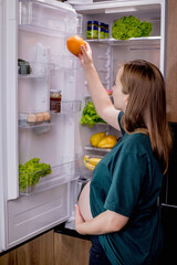 Young pregnant woman with an orange near the refrigerator at home. Healthy Eating