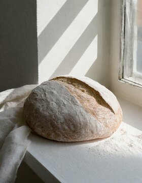 A hearty sourdough loaf with a crisp, scored crust, cooling by an open window where morning light gently illuminates its artisanal shape and the flour dusted on its surface