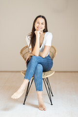 Portrait of confident beautiful woman with long brown hair, wearing casual clothes, sitting on chair in tight jeans and white t-shirt, studio background