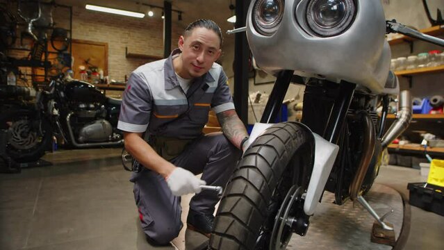 Wide shot of cheerful repairman examining wheel of motorcycle and turning to camera in garage