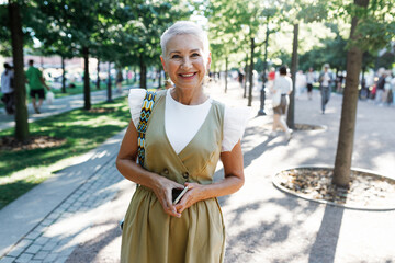 Summer walk of elegant designer wearing fashionable trendy dress outdoor at city park. Happy cheerful businesswoman of 50s having stroll on her way home through public garden or square - 774713111