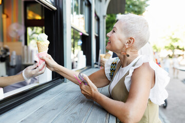 Side view of senior stylish female in trendy dress and earrings buying waffle pistachio ice cream outdoor, having rest during walk at city center on summer evening, visiting new spots - 774713124