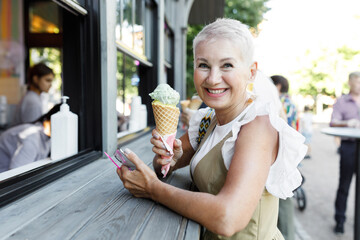 Side view of cheerful aged hipster female with short pink hair leaning on counter after buying waffle ice cream outdoor, looking at camera with candid childish smile during summer stroll in city - 774713181