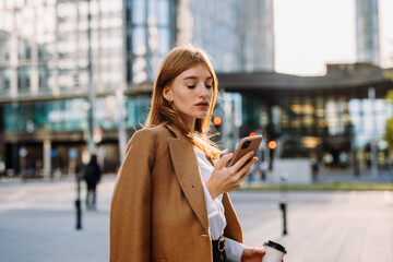 Young smiling business woman in jacket with coffee cup using mobile phone