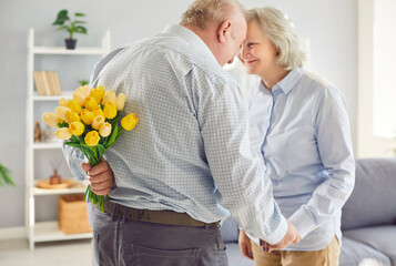 Happy senior man with tulips behind back making romantic present to his smiling wife on marriage...