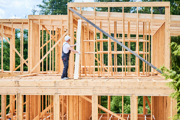 Fototapeta premium Carpenter constructs wooden-framed house. Man holds large plank in his hands while dressed in work clothes and helmet. Concept of modern ecological construction.
